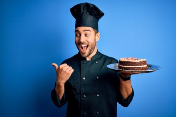 Young handsome cooker man with beard wearing uniform and hat holding tray with cake pointing and...