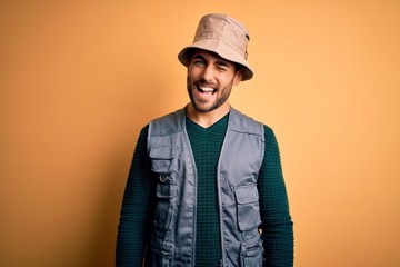 Handsome tourist man with beard on vacation wearing explorer hat over yellow background winking...