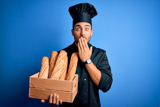 Young cooker man with beard wearing uniform holding box with bread over blue background cover mouth with hand shocked with shame for mistake, expression of fear, scared in silence, secret concept
