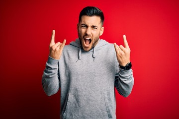 Young handsome sportsman wearing sweatshirt standing over isolated red background shouting with crazy expression doing rock symbol with hands up. Music star. Heavy concept. © Krakenimages.com