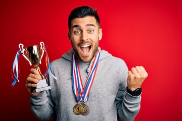 Young handsome succesful man holding trophy wearing medals over red background screaming proud and...