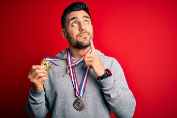 Young handsome succesful man winning medals standing over isolated red background serious face...
