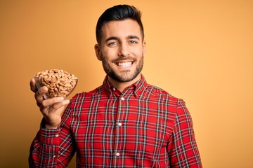 Young handsome man holding bowl with healthy peanuts over isolated yellow background with a happy face standing and smiling with a confident smile showing teeth