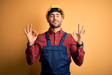Young builder man wearing construction uniform and safety helmet over yellow isolated background...