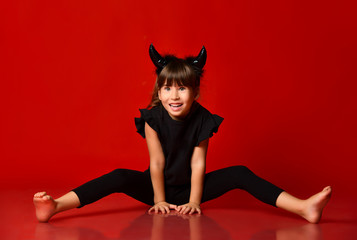 Child with devil horns, in black blouse and leggings. She is smiling, sitting on floor with legs apart. Red background. Close up