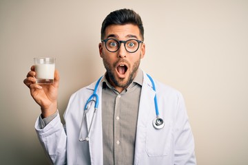 Young doctor man wearing stethoscope holding a glass of milk over isolated background scared in shock with a surprise face, afraid and excited with fear expression