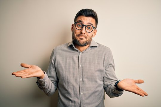 Young handsome man wearing elegant shirt and glasses over isolated white background clueless and confused expression with arms and hands raised. Doubt concept.