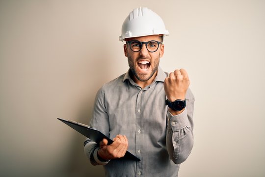 Young architect inspector man wearing builder safety helmet and using clipboard annoyed and frustrated shouting with anger, crazy and yelling with raised hand, anger concept