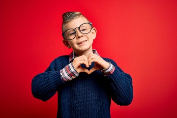 Young little caucasian kid with blue eyes standing wearing smart glasses over red background smiling in love showing heart symbol and shape with hands. Romantic concept.