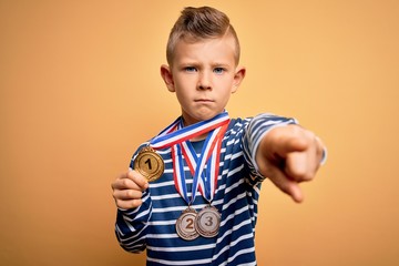 Young little caucasian winner kid wearing award competition medals over yellow background pointing with finger to the camera and to you, hand sign, positive and confident gesture from the front