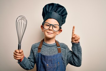 Young little caucasian cook kid wearing chef uniform and hat using manual whisk surprised with an...