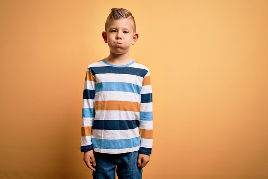 Young little caucasian kid with blue eyes wearing colorful striped shirt over yellow background puffing cheeks with funny face. Mouth inflated with air, crazy expression.