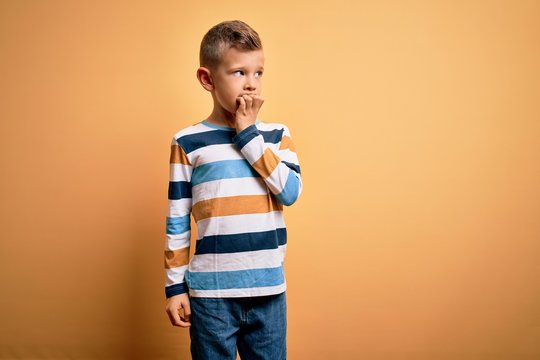 Young little caucasian kid with blue eyes wearing colorful striped shirt over yellow background looking stressed and nervous with hands on mouth biting nails. Anxiety problem.