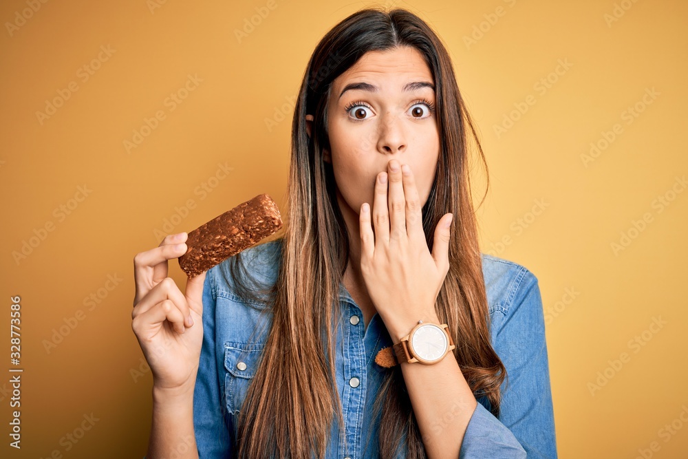 Sticker Young beautiful girl holding healthy protein bar standing over isolated yellow background cover mouth with hand shocked with shame for mistake, expression of fear, scared in silence, secret concept