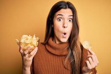 Young beautiful girl holding bowl with chips potatoes standing over yellow background scared in...