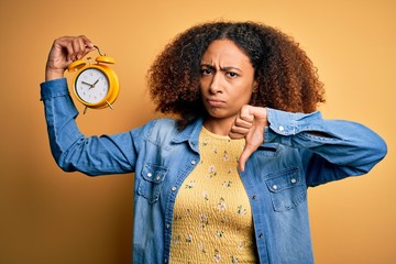 Young african american woman with afro hair holding vintage alarm clock over yellow background with...