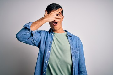 Young handsome man wearing casual shirt and glasses over isolated white background peeking in shock covering face and eyes with hand, looking through fingers with embarrassed expression.