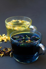 Hot butterfly pea tea and Jasmine tea. Healthy lifestyle. glass cups of blue and yellow herbal tea on a black background. Hot drinks. two cups of hot tea with dry flowers on a black table. vertical
