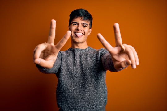Young handsome man wearing casual sweater standing over isolated orange background smiling with tongue out showing fingers of both hands doing victory sign. Number two.