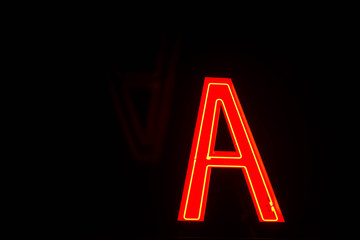 Neon red letter A with a faint upside down reflection in the black background. A neon sign at night.