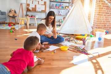 Young beautiful teacher and toddlers sitting on the floor drawing around lots of toys at kindergarten