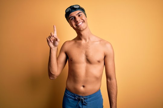 Young handsome man shirtless wearing swimsuit and swim cap over isolated yellow background pointing finger up with successful idea. Exited and happy. Number one.