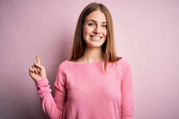 Young beautiful redhead woman wearing casual sweater over isolated blue background with a big smile on face, pointing with hand finger to the side looking at the camera.