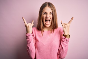 Young beautiful redhead woman wearing casual sweater over isolated pink background shouting with crazy expression doing rock symbol with hands up. Music star. Heavy music concept.