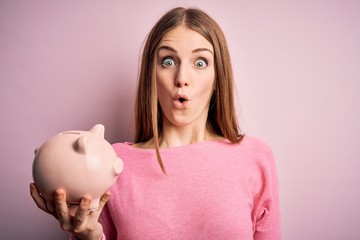 Young beautiful redhead woman holding piggy bank over isolated pink background scared in shock with...