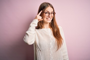 Young beautiful redhead woman wearing casual sweater and glasses over pink background Smiling pointing to head with one finger, great idea or thought, good memory