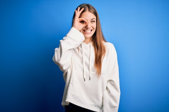 Young beautiful redhead sporty woman wearing sweatshirt over isolated blue background doing ok gesture with hand smiling, eye looking through fingers with happy face.