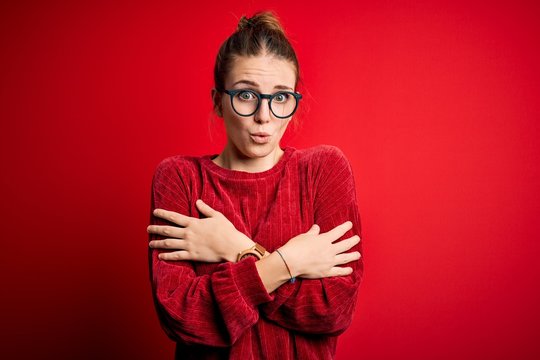 Young beautiful redhead woman wearing casual sweater over isolated red background shaking and freezing for winter cold with sad and shock expression on face