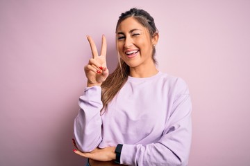 Young beautiful sport woman wearing sweatshirt over pink isolated background smiling with happy face winking at the camera doing victory sign. Number two.