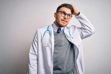 Young doctor man with blue eyes wearing medical coat and stethoscope over isolated background smiling confident touching hair with hand up gesture, posing attractive and fashionable