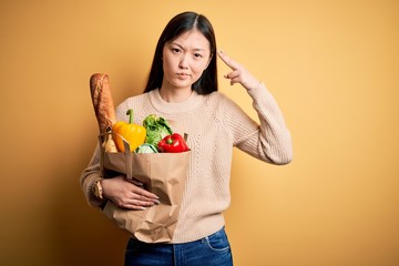 Young asian woman holding paper bag of fresh healthy groceries over yellow isolated background Shooting and killing oneself pointing hand and fingers to head like gun, suicide gesture.