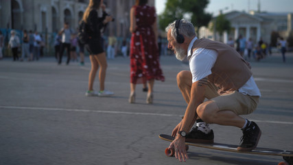The light-skinned hipster skates and listens to music outdoors. Intelligent middle-aged man rides a longboard and looks forward.