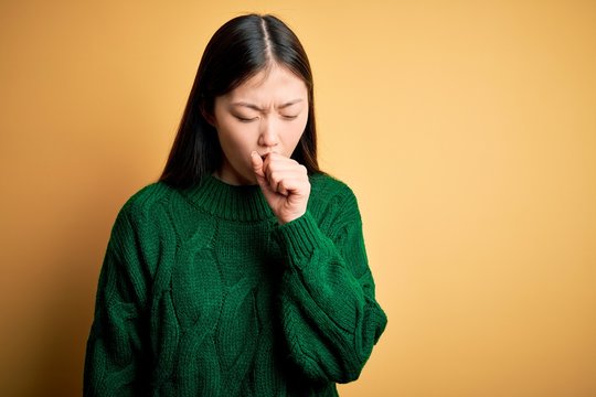 Young beautiful asian woman wearing green winter sweater over yellow isolated background feeling unwell and coughing as symptom for cold or bronchitis. Health care concept.
