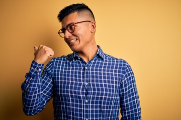 Young handsome latin man wearing casual shirt and glasses over yellow background smiling with happy face looking and pointing to the side with thumb up.