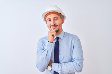 Young business man wearing contractor safety helmet over isolated background looking confident at the camera smiling with crossed arms and hand raised on chin. Thinking positive.