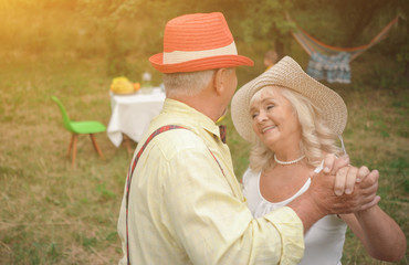 The Middle-aged Man And Lady Are Dancing In The Beautiful Garden.The Charming Lady Is Looking At His Husband With Love And Admiration.Cosy Hammock And Table With Chairs Are Behind Them