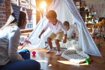 Beautiful teacher and blonde toddler twins playing inside tipi around lots of toys at kindergarten