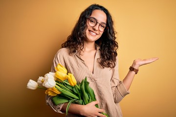 Young beautiful romantic woman with curly hair holding bouquet of yellow tulips smiling cheerful presenting and pointing with palm of hand looking at the camera.