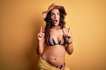Young beautiful woman with curly hair on vacation wearing bikini and summer hat amazed and surprised looking up and pointing with fingers and raised arms.