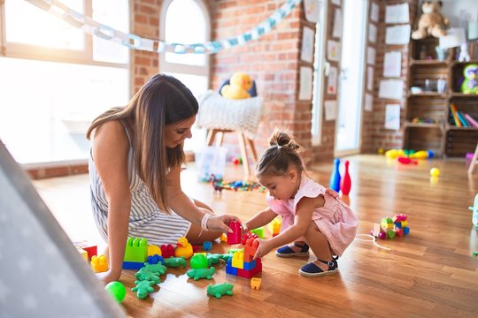 Young beautiful teacher and toddler sitting on the floor playing with building blocks toy at kindergarten