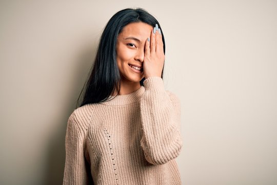Young beautiful chinese woman wearing casual sweater over isolated white background covering one eye with hand, confident smile on face and surprise emotion.