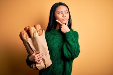 Young beautiful chinese woman holding paper bag with bread over isolated yellow background serious...