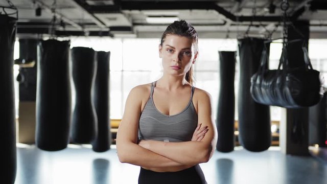 Tilt down waist up shot of serious fit young woman walking towards camera and posing cross armed looking at camera in boxing gym with heavy bags in background