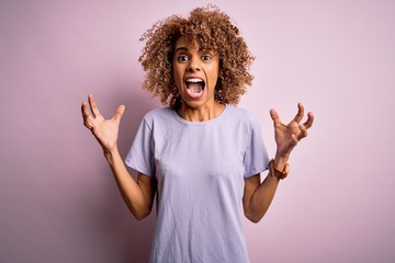 Young beautiful african american woman wearing casual t-shirt standing over pink background crazy and mad shouting and yelling with aggressive expression and arms raised. Frustration concept.