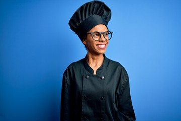Young african american chef woman wearing cooker uniform and hat over blue background looking away to side with smile on face, natural expression. Laughing confident.