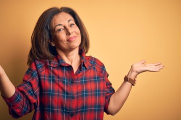 Middle age beautiful woman wearing casual shirt standing over isolated yellow background clueless and confused expression with arms and hands raised. Doubt concept.